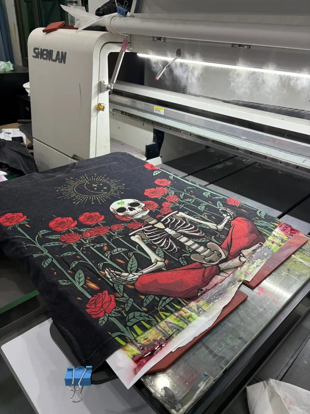 Large skeleton and red rose graphic on a dark T-shirt positioned under a garment printer